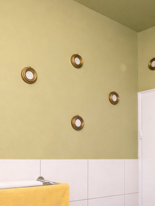 Hellgrüne Badezimmerwand mit mehreren kleinen, runden Spiegeln in goldfarbenen Rahmen, unregelmäßig angeordnet. Unterhalb der Wand befinden sich weiße Fliesen, rechts ist eine weiße Tür zu sehen, im Vordergrund hängt ein gelbes Handtuch an einer Metallstange.