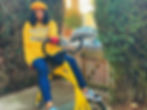 Roxy, a non-binary person of colour, wears a yellow jumper that reads rollin with the homies, with a matching yellow beret and blue jeans. They sit on their matching yellow bike which is specialised for standing.