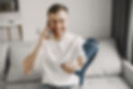 Sitting on a white sofa, a white man with brown hair wears a white t-shirt and round glasses. He holds his phone to one ear, while holding his crutch in front of him with the other.