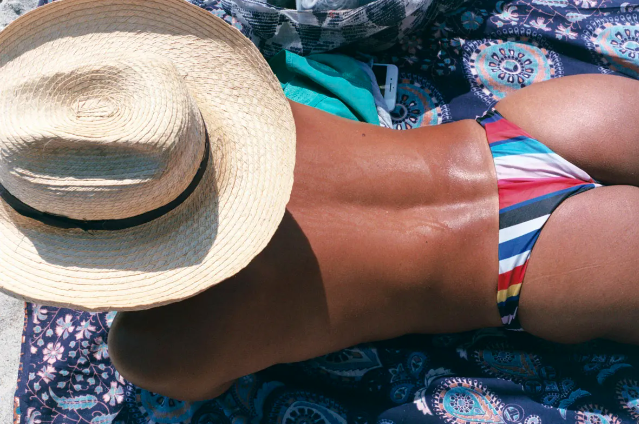 This photo is a close crop of someone lying on a patterned towel on the beach. Their head is not visible under a wide brimmed straw hat, they are wearing striped speedos and their back has visibly tanned. 