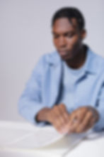 Against a grey background, a black man with dark hair in short braids, wearing a light blue jacket and matching t-shirt sits at a table. His eyes are down, as he touches a book that is printed in Braille.