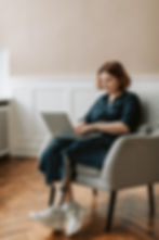 A white woman with short red hair is wearing a dark green dress and white trainers. She is sitting in a white armchair, typing on a laptop that sits on her knee. She has one prosthetic leg, which is crossed over her other leg at the ankles. In the background there is a beige wall with white wooden panelling and a dark wooden floor.