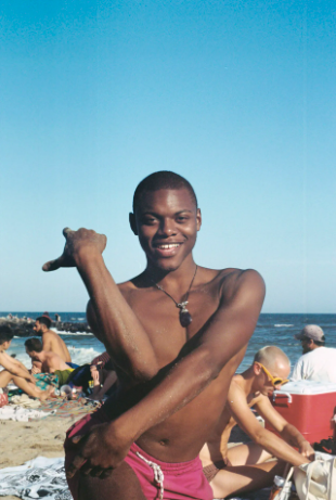 This photo is of a black person with buzzed hair, they are shirtless and wearing pink swimming trunks. They are smiling at the camera and in the midst of voguing on the beach. Behind them you can see other beachgoers and the sea in the distance.