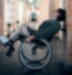 The image shows a white woman in a manual wheelchair with short dark hair with her laptop on her knee. She is wearing black glasses, a dark green blazer, beige trousers and white trainers. She is in an office, and has pivoted her chair backwards so the front wheels are off the floor. She leans her head against the brick wall behind her.