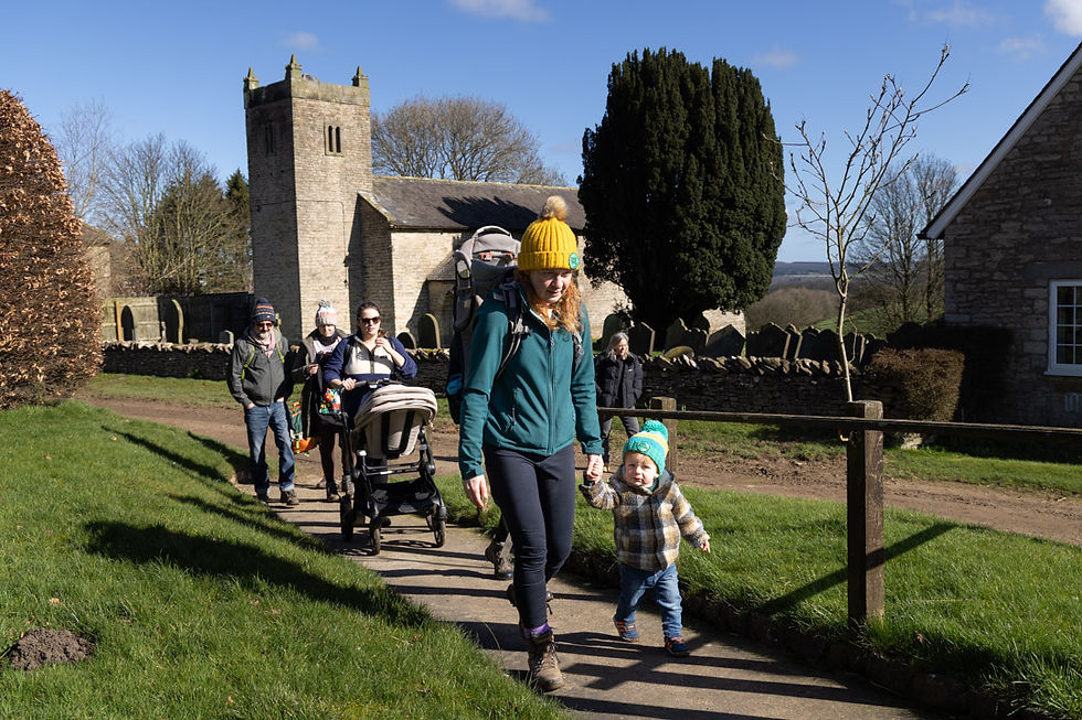 “A family with a young child and other pilgrims, including a person pushing a buggy, walk past Saint Michael’s Church during the Saint Aelred’s Pilgrim Trail pilgrimage.”