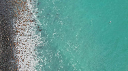 Bird's-eye view of two surfers at Magnet Bay, one paddling toward an exciting wave, rocky coastline, and crisp aqua-blue waters.