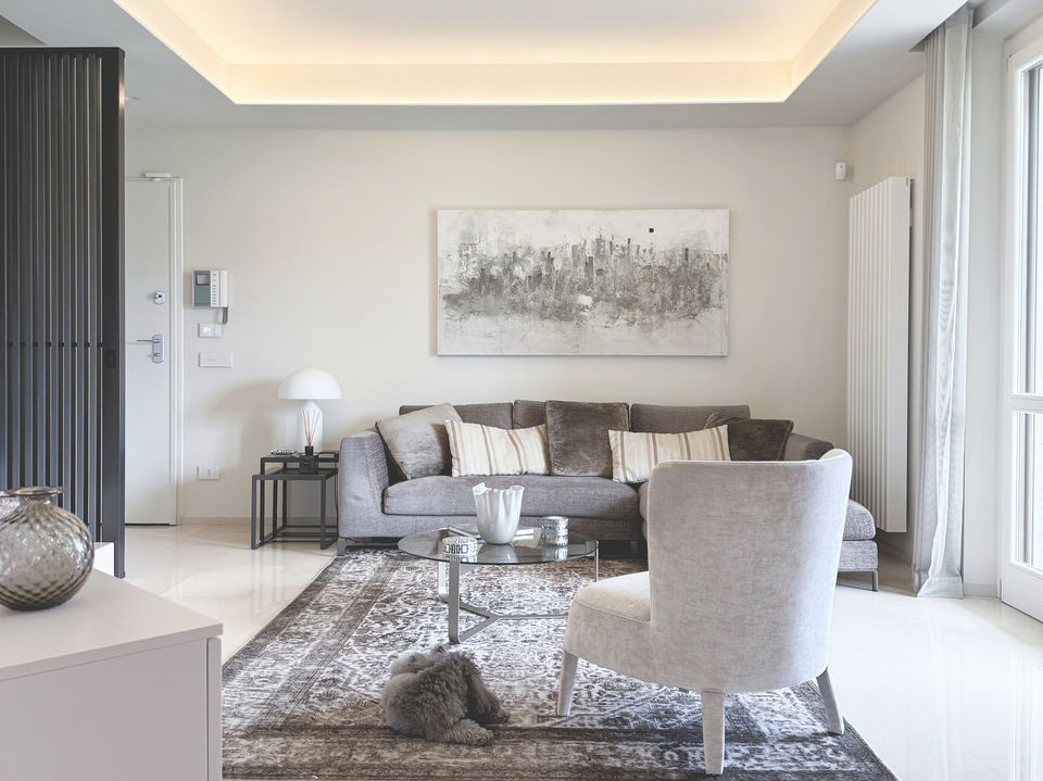 Area living ristrutturata con divano, poltrona, quadro astratto, tappeto e pannellatura in legno scuro, toni grigi e beige.