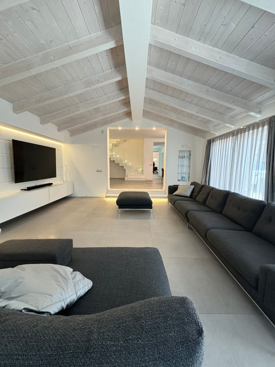 Living minimalista in villetta indipendente con divano grigio, soffitto in legno chiaro a vista e scala in marmo illuminata.
