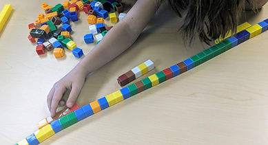 a child's hand holds one small stick of unisex cubes against a much larger stick of unisex cubes. Other unified cubes are scattered on the table in the background