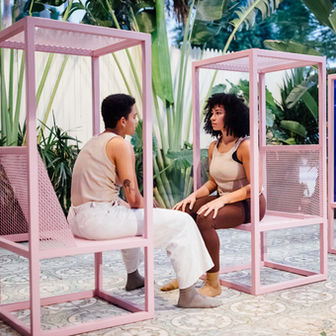 two people sit facing one another each sitting on a seat inside of a rectangular pink steel cube