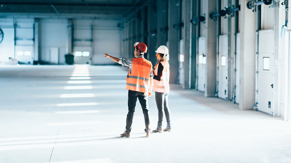 Two people in safety gear and orange vests, standing in a large industrial space. One points forward. Bright sunlight and shadows across the floor.