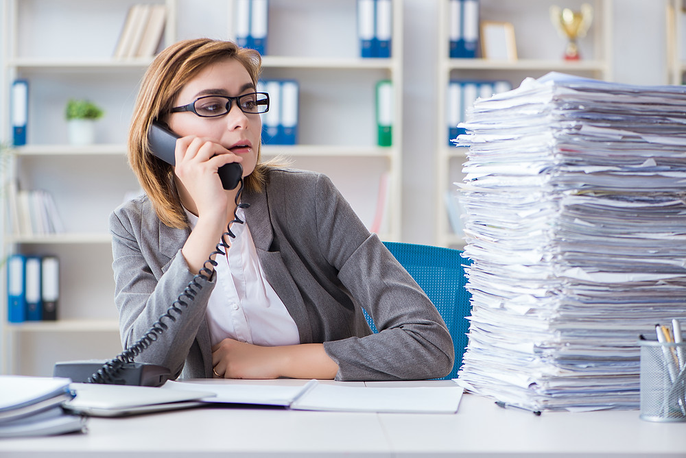 Insurance staff member on the phone while staring at the large stack of paperwork she still has to do.