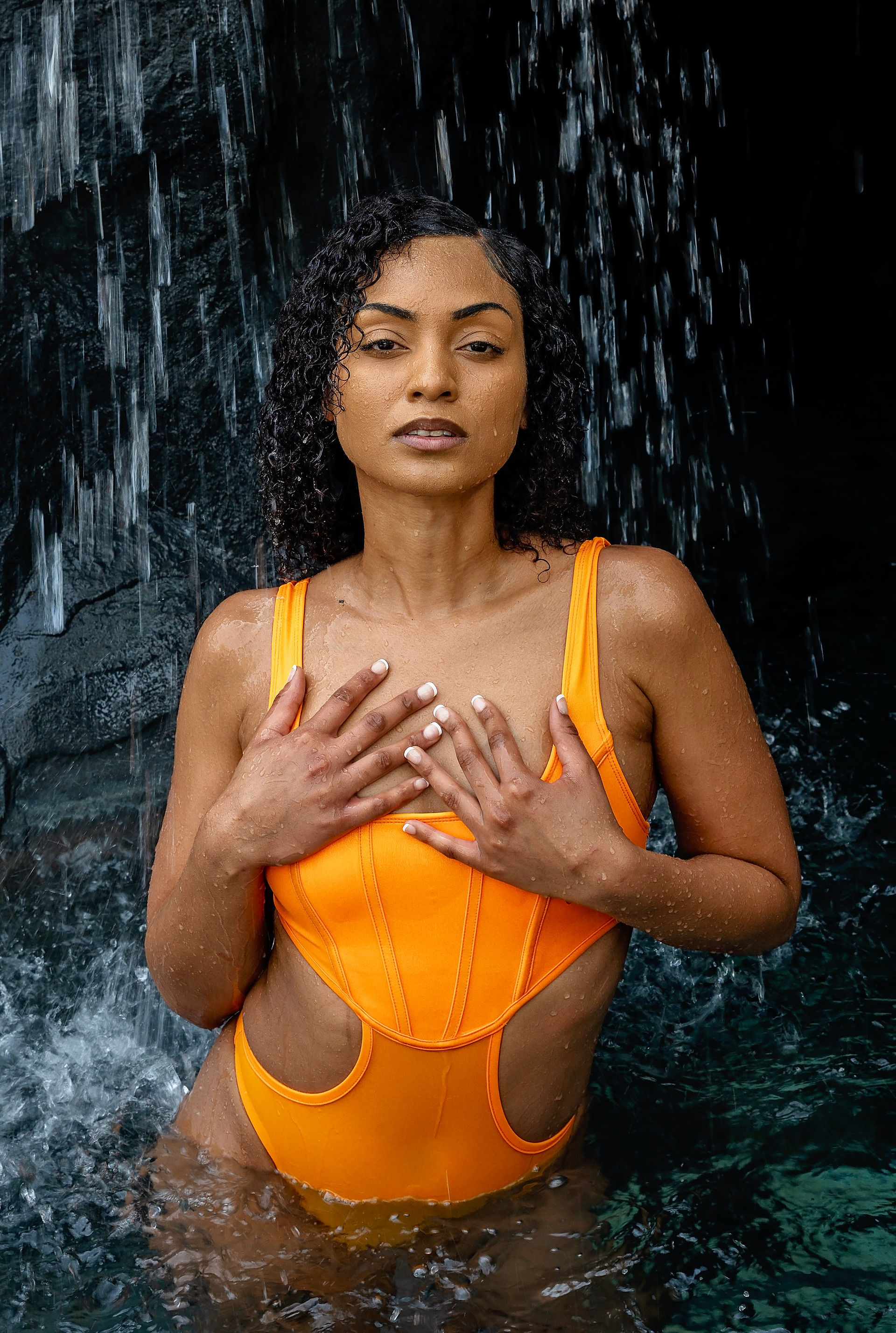 Woman in orange swimsuit posing against a dark waterfall background.