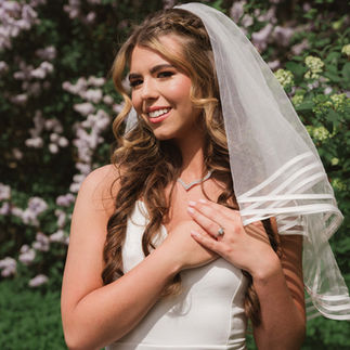 Smiling bride in white dress, veil, standing among purple flowers.