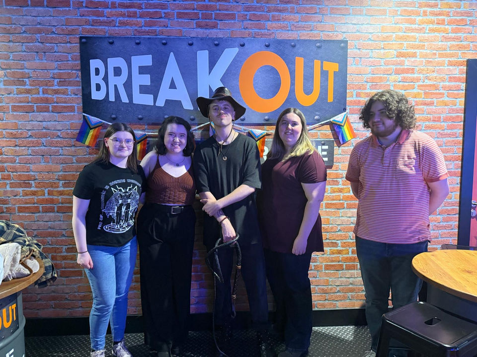 Group photo in front of BREAKOUT. Five people smiling by the sign BREAKOUT ROOMS.