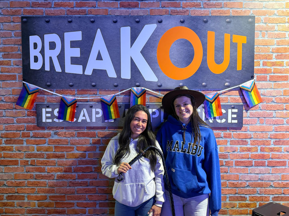 Two women smiling near Breakout Escape Place sign, surrounded by Pride flags.