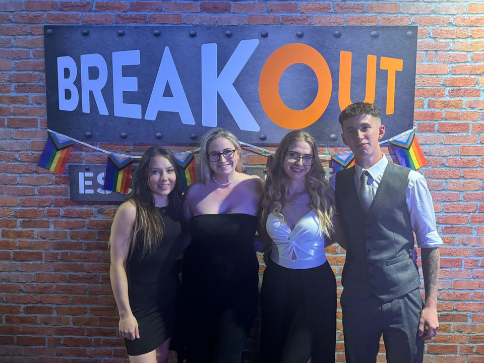 Four people pose in front of the Breakout sign with pride flags