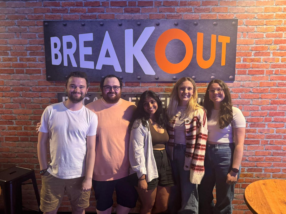 Group poses under Breakout sign, brick background. BREAKOUT ROOMS, team smiles.