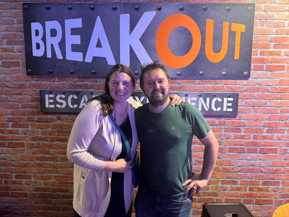 Two people pose in front of a BREAKOUT ESCAPE room sign.