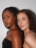 Two women stand back-to-back against a gray background, with natural expressions. One has braided hair, the other loose with clips. Both wear black tops. Skin- focused makeup.