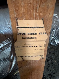 Torn, aged label on wooden beam reads “STOS FIBER FLAKE Insulation.” Surroundings include silver ductwork and dark textured floor. Asbestos inspection in Colorado
