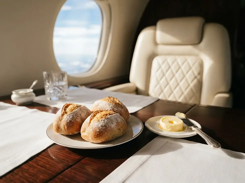 Three bread rolls and butter on a polished airplane table by a window, with a glass and bright sky outside.