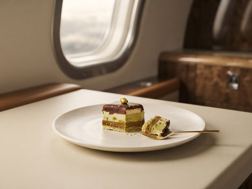 Slice of pistachio layered cake with chocolate glaze on a white plate and gold fork beside an airplane window.