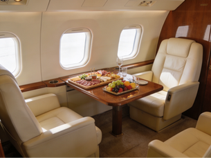 Plush leather seats in a private jet with a wooden table set for a feast, featuring fruits and charcuterie, against bright windows. Luxurious setting.