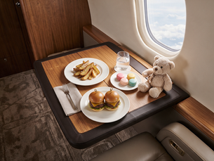 Private jet table with on top burgers and fries, macarons, a glass of water, a fork and a light brown teddy bear.