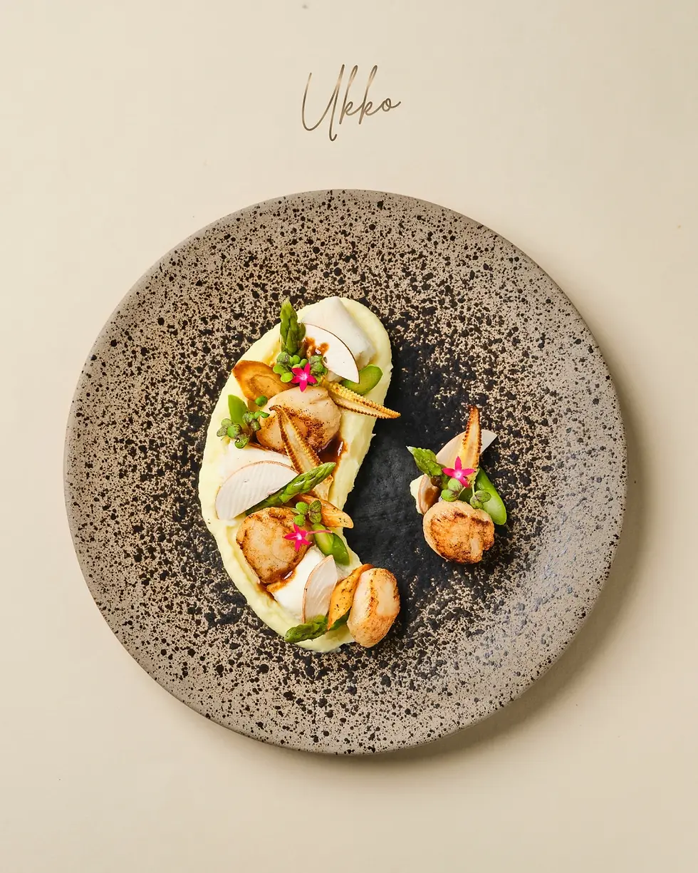Gourmet scallop dish on speckled black plate, garnished with asparagus and pink flowers, with Ukko above on a cream background