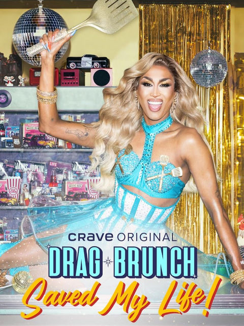 A drag queen is dressed in a bright blue dress with mini silver spatulas fashioned in an X over each breast. She holds a large spatula above her head as she smiles at the camera, mouth wide.