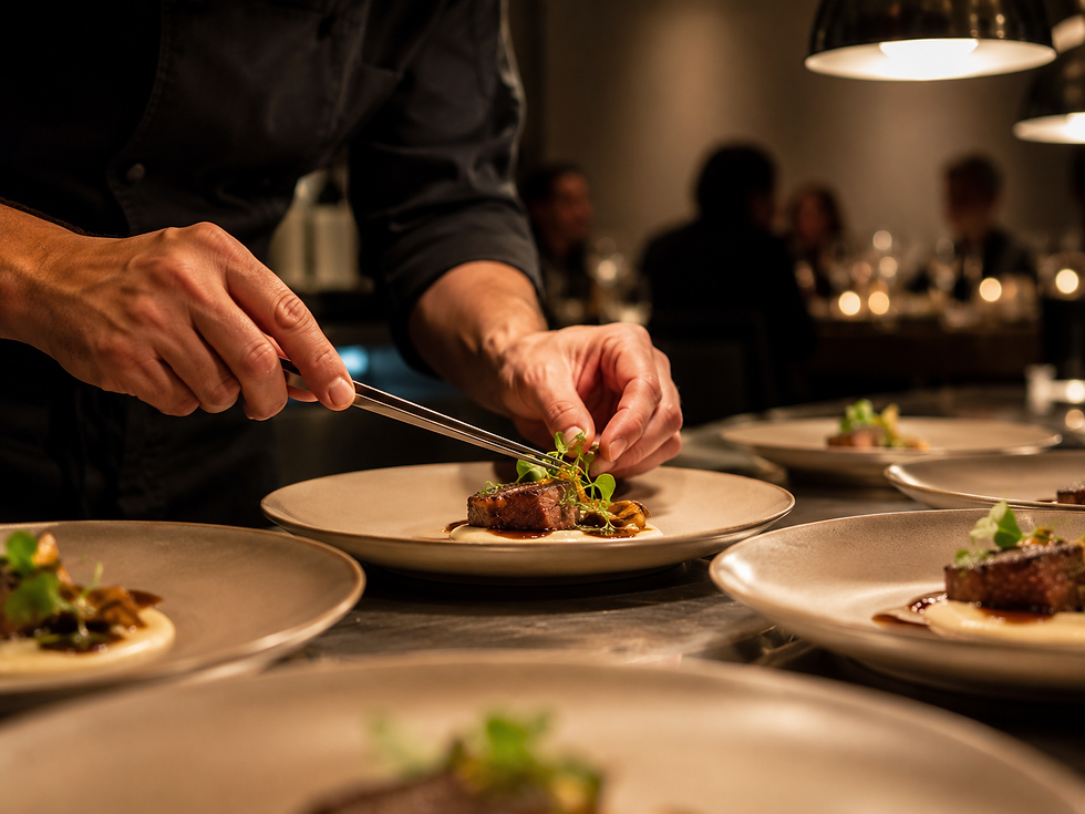 A chef finishes several plates moments before service, while guests wait in the softly lit dining room beyond.