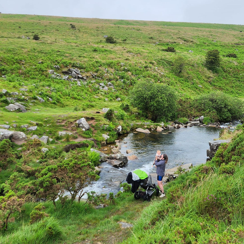 Person with baby in a green landscape near a small river. Stroller by their side. Overcast sky and lush grassy hills in the background.