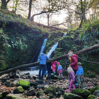 A group of adults and children hike uphill in a grassy, wooded area. They wear colourful jackets, and the mood is cheerful under a sunny sky.