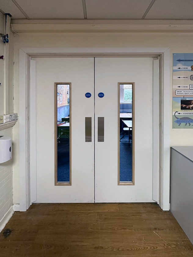 A set of white double doors with narrow vertical windows near the center, located in an indoor setting with wooden flooring and visible blue "Fire Door Keep Shut" signs.