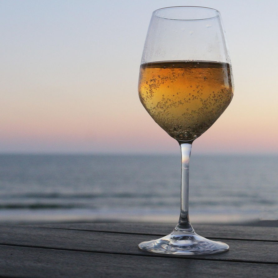 A champagne glass with bubbles sits on a wooden table overlooking a calm ocean at sunset, with a gradient sky of soft pink and blue.