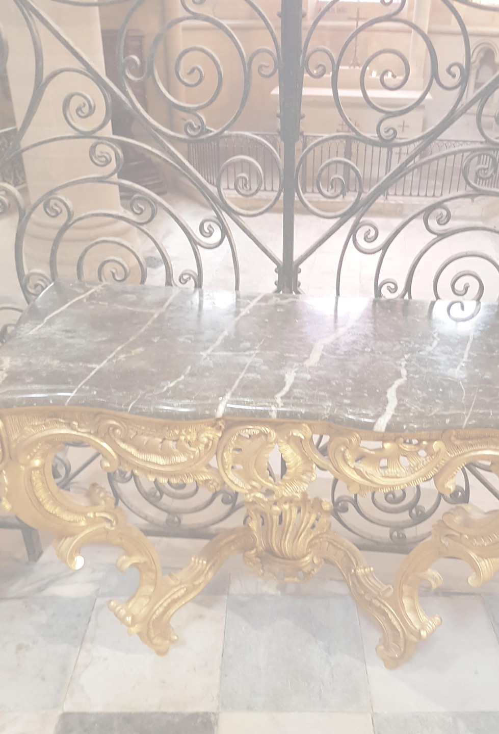 – cours de l’art - Cette superbe image nous plonge au coeur d'une majestueuse cathédrale. On découvre en premier plan une magnifique console en bois doré datant de l'époque Louis XV. Ses lignes courbes et ses délicates sculptures florales dans le plus pur style Rococo témoignent du raffinement ornemental du 18ème siècle. La console est adossée à une grille en fer forgé aux motifs très travaillés. En arrière-plan, on distingue la nef centrale de la cathédrale, baignée d'une douce lumière tamisée. L'architecture gothique se dévoile à travers les hauts piliers reliés par des arcs-boutants et les voûtes en ogives. En levant les yeux, on aperçoit les magnifiques vitraux qui illuminent l'édifice de mille feux. L'image offre un savant mélange des genres entre les styles Louis XV et gothique. La console rocaille contraste avec la pierre brute de la cathédrale. Cette photo nous transporte au coeur de l'art et de l'architecture des 17ème et 18ème siècles.