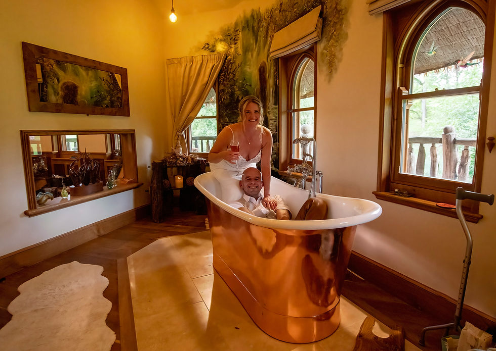 Smiling couple in a copper bathtub with champagne in a warm, rustic bathroom with arched windows and forest view.