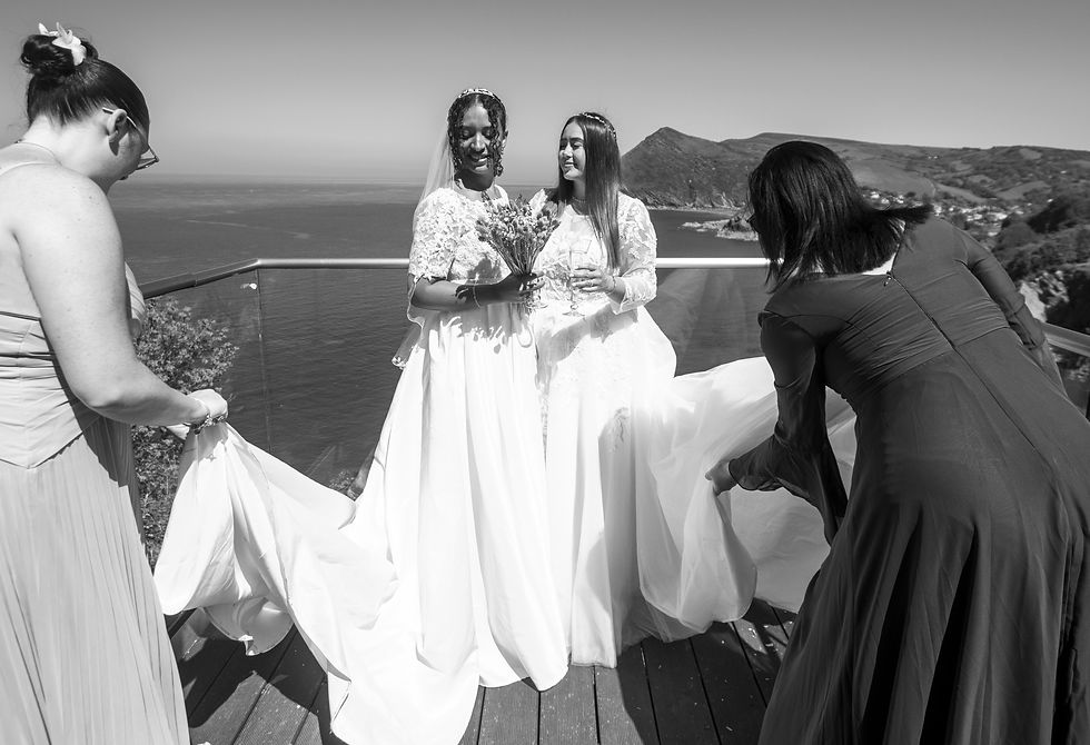 Two bridesmaids wafting the dresses of two brides as they prepare to be photographed.