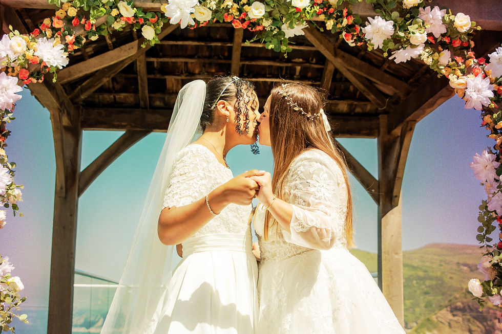 Two brides kiss under a floral arch. Both wear white dresses and hold hands. The setting is outdoor with clear blue skies. Romantic mood.