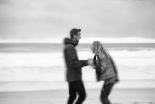 Older couple walking hand in hand along a wintery beach, laughing and enjoying each other’s company as cold waves and soft gray skies frame the peaceful scene.