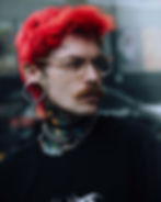 Man with bright red hair looks to the side wearing jewelry - including the vampire the masquerade choker, with the black dice