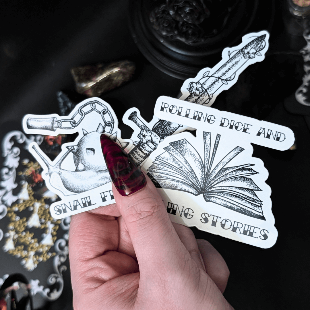 Hand holding stickers: snail flail, a vine wrapped sword, and a book with "rolling dice and telling stories" around it