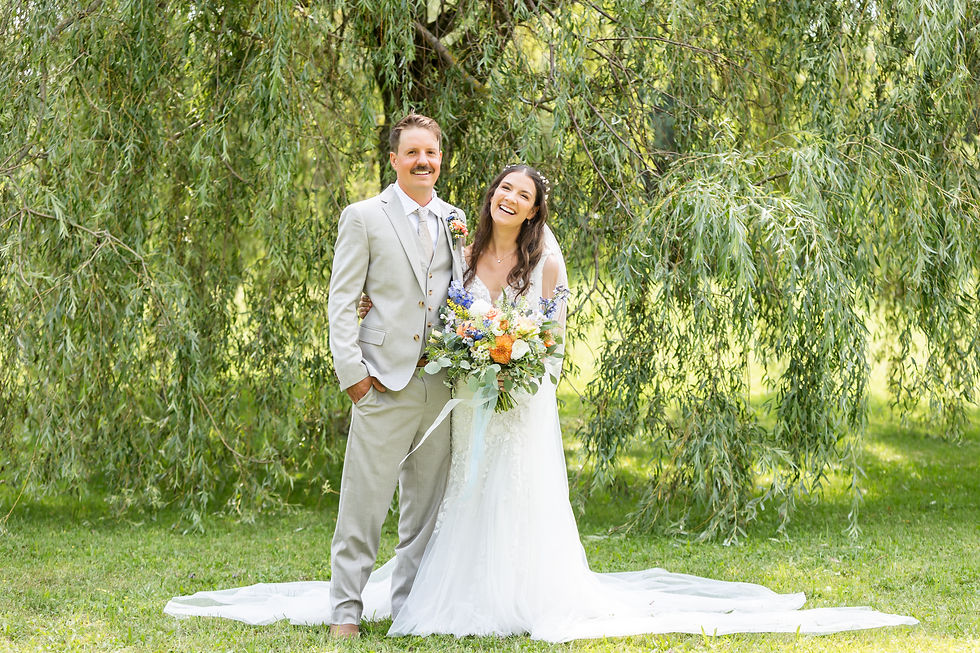 Smiling bride and groom stand under drooping green trees, bride holding colorful bouquet, in a sunny outdoor wedding portrait in Fergus, Ontario.