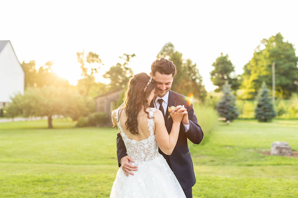 Bride and groom slow dance in a sunny garden at West Avenue Cider in Freelton, smiling warmly; her lace dress and his dark suit glow in golden light.