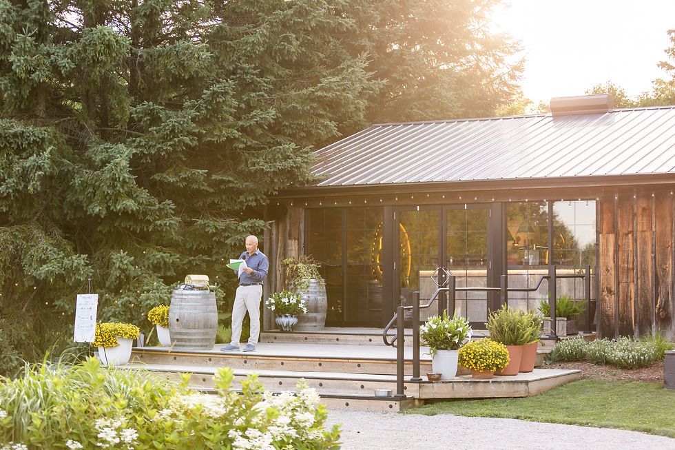A man reads papers on a wooden deck outside a rustic building surrounded by potted plants and trees, bathed in warm sunlight. Wedding at West Avenue Cider House. Photos taken by Kayla Beth Photography a Guelph Wedding Photographer.