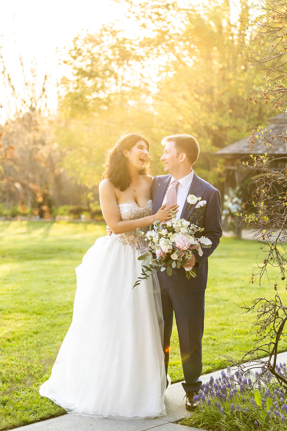 Smiling bride and groom embrace in a sunny garden at sunset at The Arboretum in Guelph, Ontario. The bride holding a pastel bouquet, warm and joyful mood.