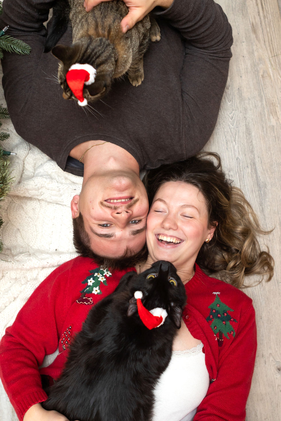 A smiling couple lies on the floor with two cats wearing Santa hats in Linwood, Ontario. Festive sweaters and cozy blankets set a joyful holiday mood.
