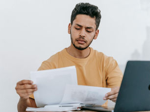 Man in a tan shirt reviews papers at a desk with a laptop, focused expression, white background. Mood is concentrated and thoughtful.