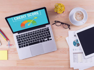 Laptop on desk displaying "Credit Score" with gauge; papers, glasses, tablet, coffee, pencils, and yellow plant surround. Bright, organised setting.
